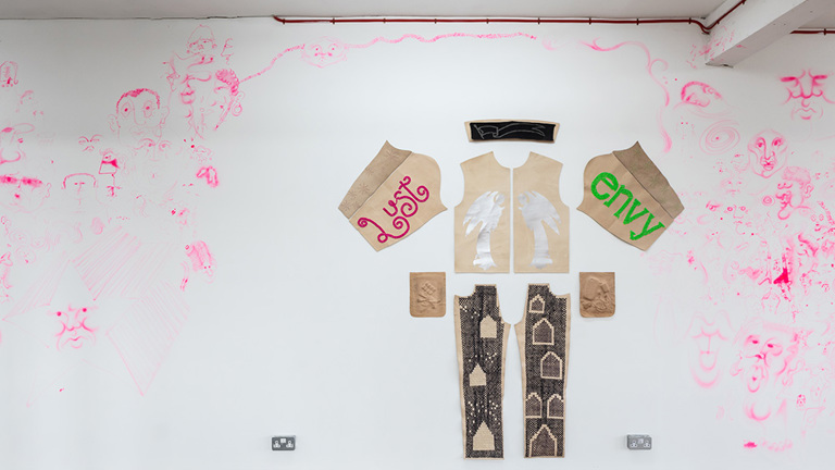 A pattern of a shirt and trousers is laid flat on a wall, decorated with art including the words 'lust' and 'envy' . On the white wall around the piece are drawings in pink.