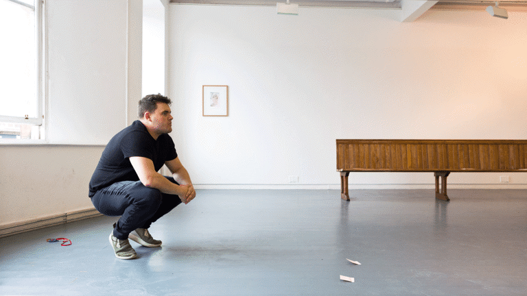 a caucasian person with short brown hair and stubble crouches in a gallery. 