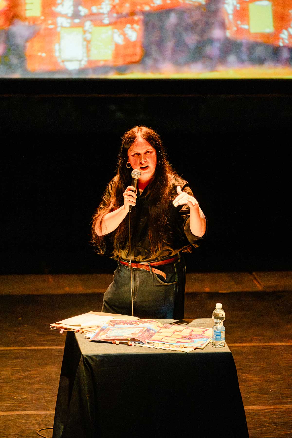 A performer speaks into a microphone behind a table covered with papers and an open illustrated book during a stage presentation.