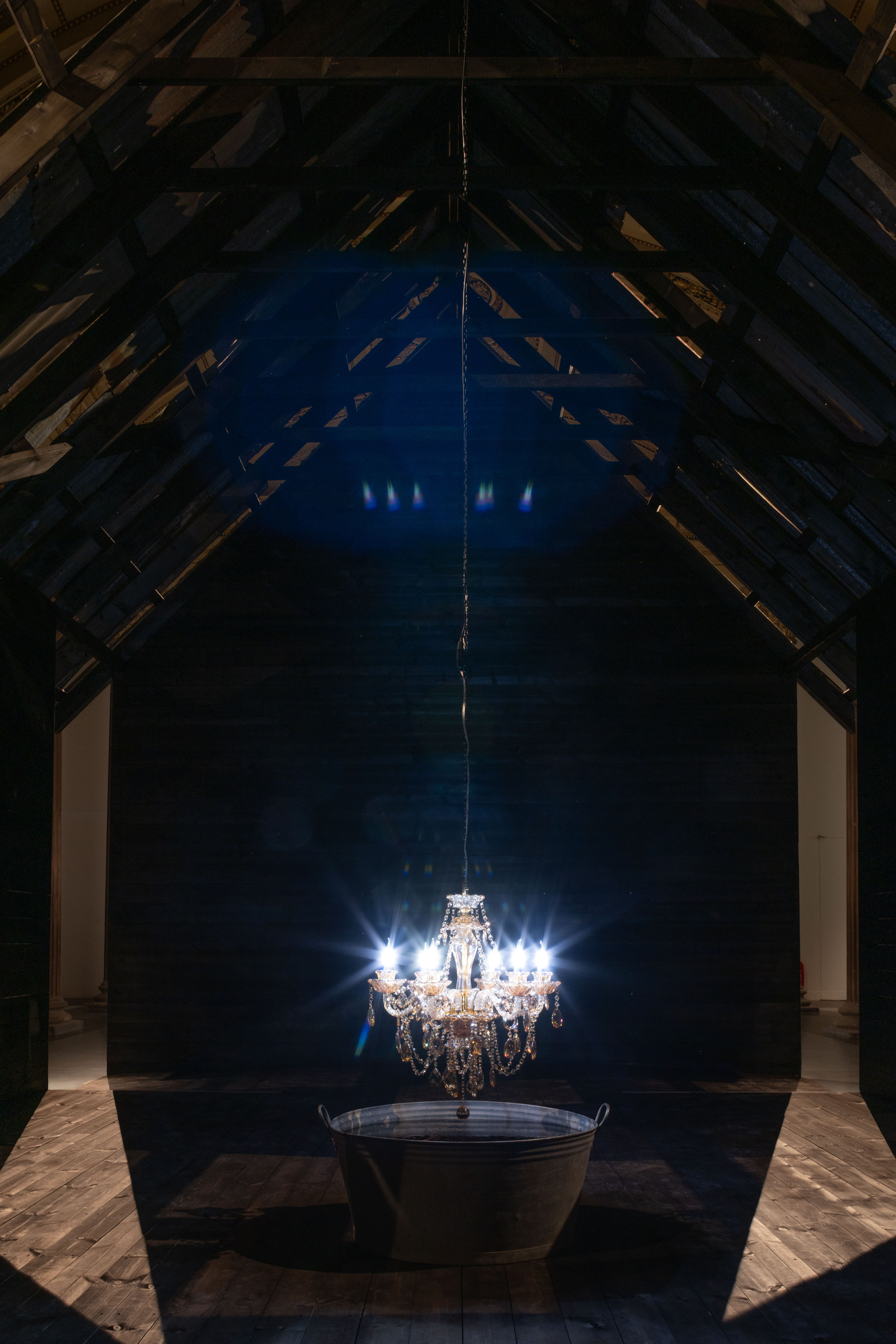 Installation view inside a dark timber-framed structure with a peaked roof, a lit crystal chandelier suspended low over a large metal wash tub in the centre of the space.