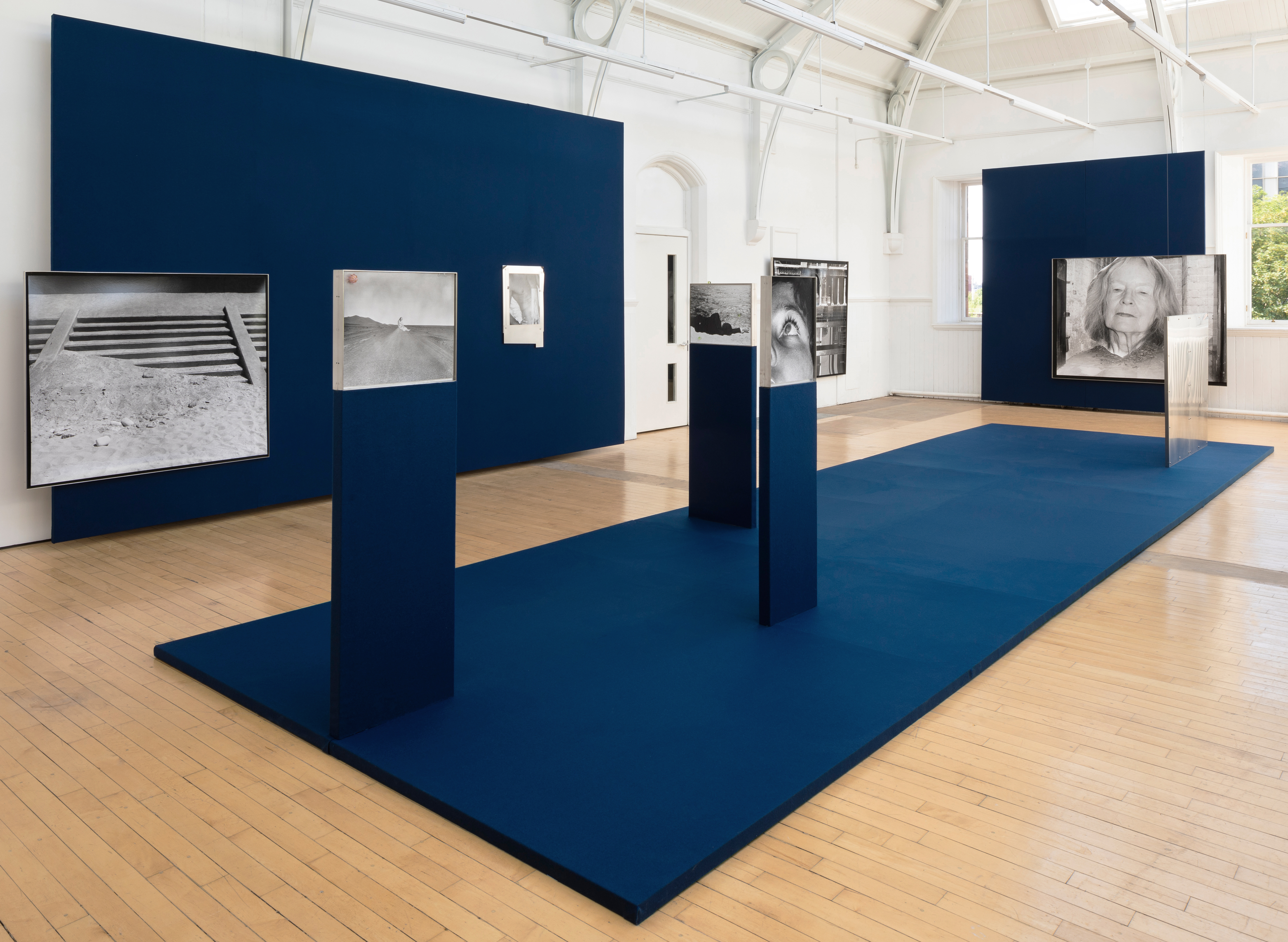 A gallery installation with navy blue walls and a raised navy platform, displaying a series of black and white photographs on plinths and walls, including a desert road, an eye, and an elderly woman's portrait, in a light-filled hall with an arched ceiling.