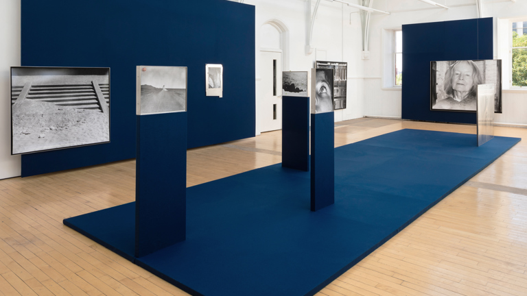 A gallery installation with navy blue walls and a raised navy platform, displaying a series of black and white photographs on plinths and walls, including a desert road, an eye, and an elderly woman's portrait, in a light-filled hall with an arched ceiling.