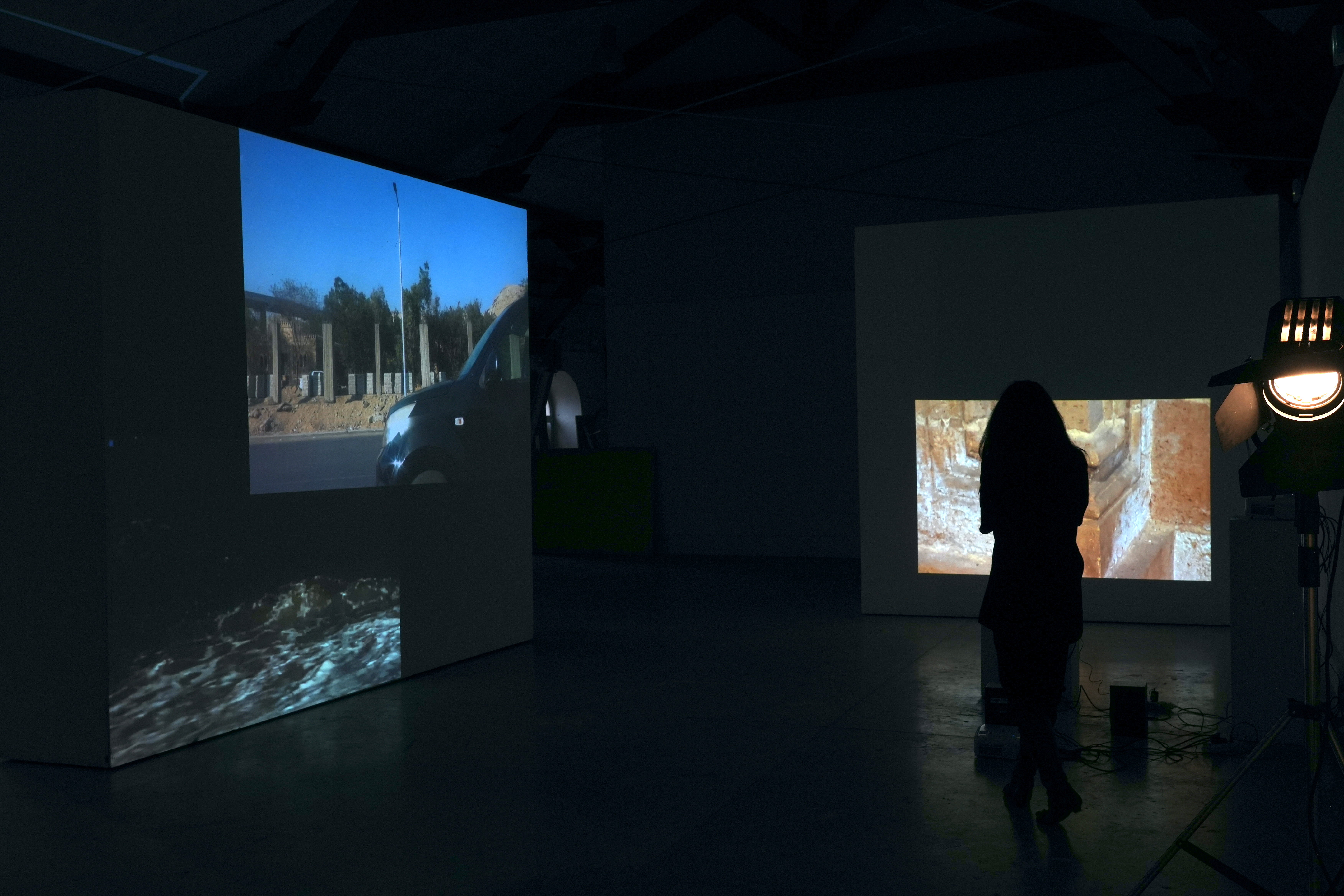 The silhoutte of a long-haired figure stands in a dark room before two project screens. The screens display three images: the ocean, a car driving past gravestones, and a close-up detail of a gravestone. 