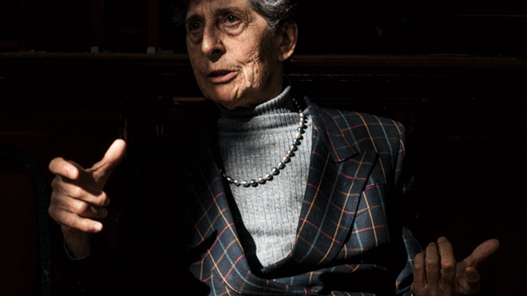  With a spotlight in her face, Esther Ferrer, white woman in her 80s with grey short hair, is talking and gesticulating with her hands. She wears a grey sweater, a pearl necklace and a tartan black jacket with yellow, blue and pink lines. 