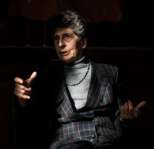 With a spotlight in her face, Esther Ferrer, white woman in her 80s with grey short hair, is talking and gesticulating with her hands. She wears a grey sweater, a pearl necklace and a tartan black jacket with yellow, blue and pink lines.