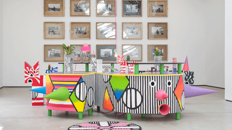 A white-walled gallery with a grid of black and white photographs on the wall behind a some multi-coloured sculptural objects in the foreground