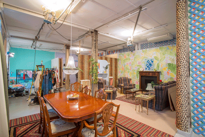 Installation view of brightly coloured interior with dining table and chairs in foreground, armchairs around a fireplace, different coloured patterned wall paper.