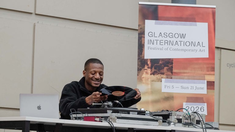 Ashley Holmes smiles as he places a vinyl record onto a turntable at a table with a laptop and DJ mixer. A Glasgow International festival banner stands behind him, listing the dates 5 to 21 June.
