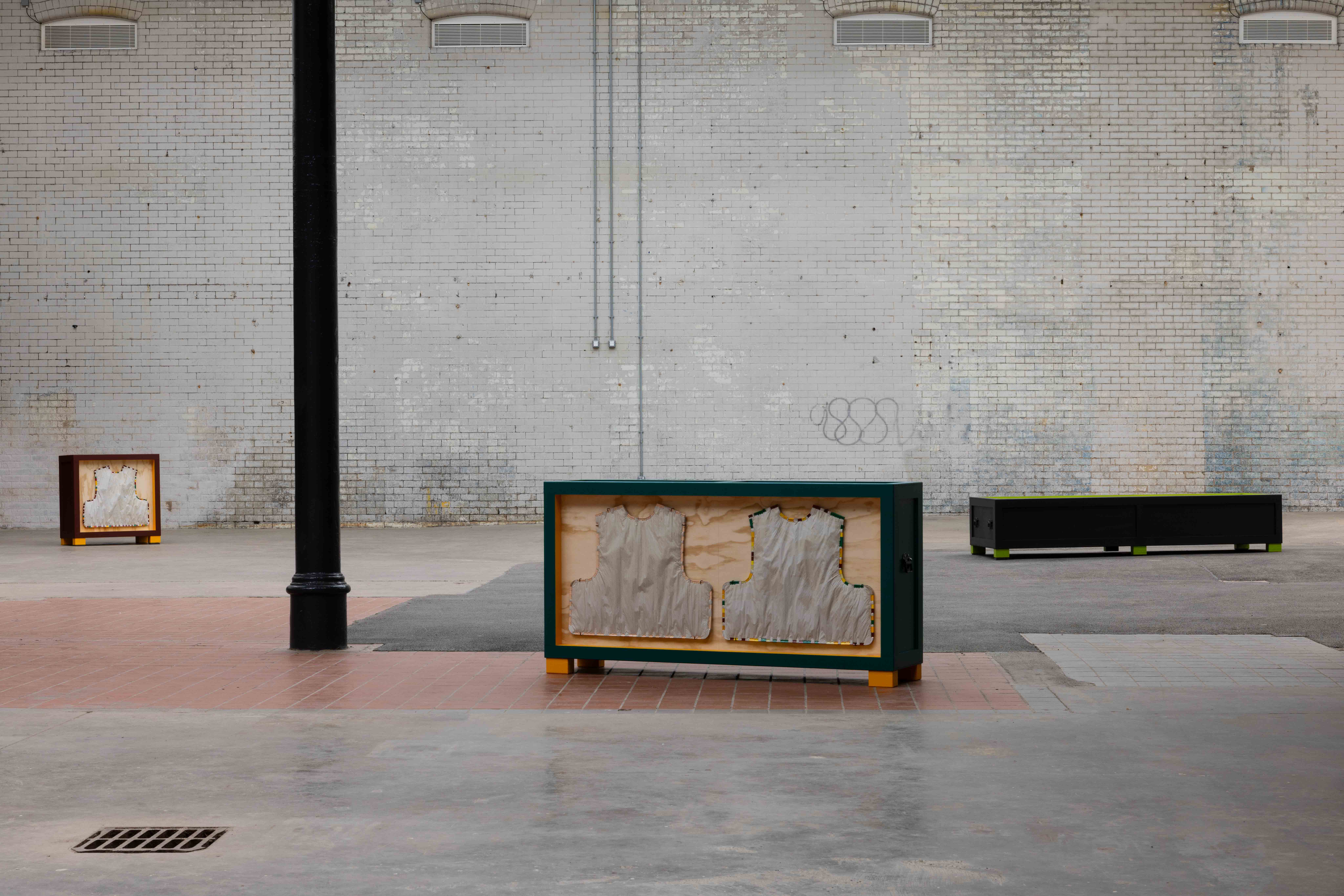 Installation view against a whitewashed brick wall, featuring three painted wooden crate-like sculptures with vest-shaped fabric forms mounted inside, spaced across a paved courtyard.