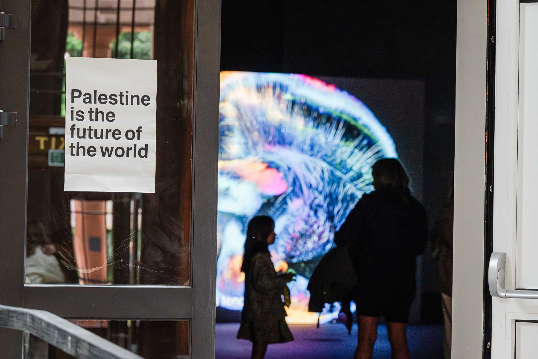 Doorway with a printed sign reading "Palestine is the future of the world" beside a gallery entrance, with two visitors silhouetted against a large colourful abstract video projection inside.