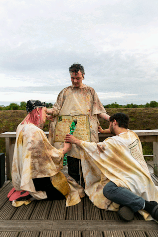 Three people wearing off white cloaks group together. One person is standing up while two other people kneel in front of them, resting a wooden pole against their cloak.