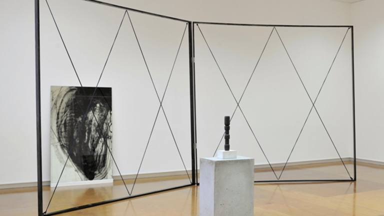 sculptural installation in a gallery space of a criss-crossing structure in a frame. in front there is a plinth with a small black sculpture. behind there is an expressive painting leaning against the wall.