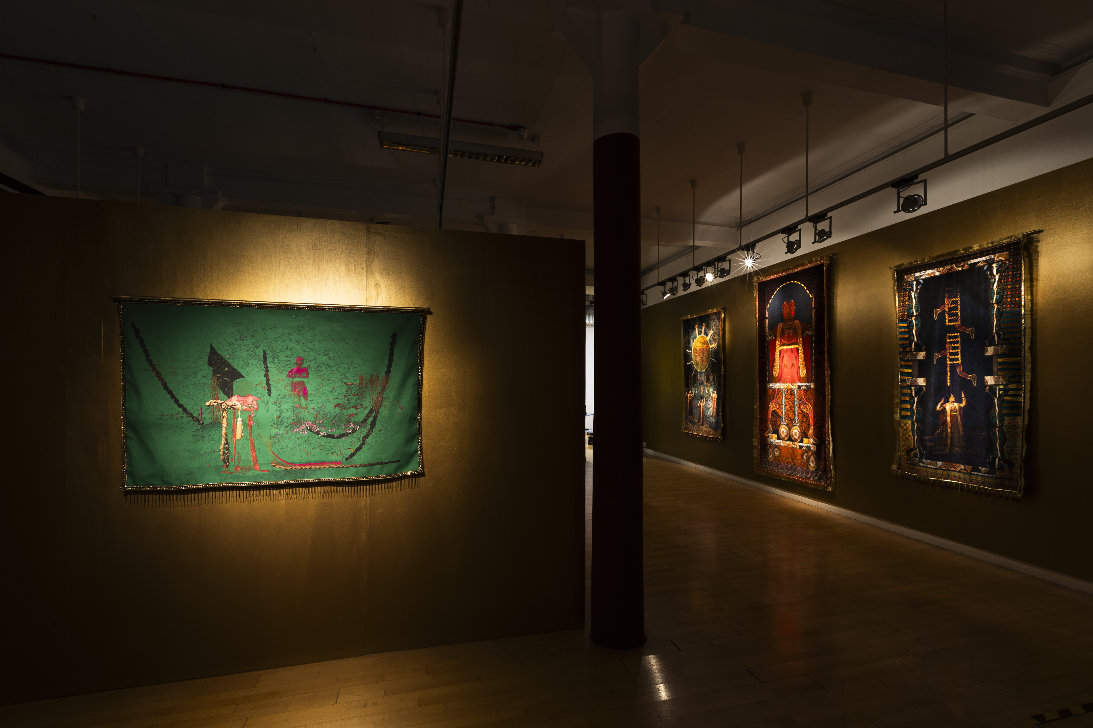 A dimly lit gallery at Streetlevel Photoworks with a green tapestry by Aqsa Arif hung on a side wall, showing a robed figure amid foliage, with three more richly coloured tapestries visible further along the corridor.