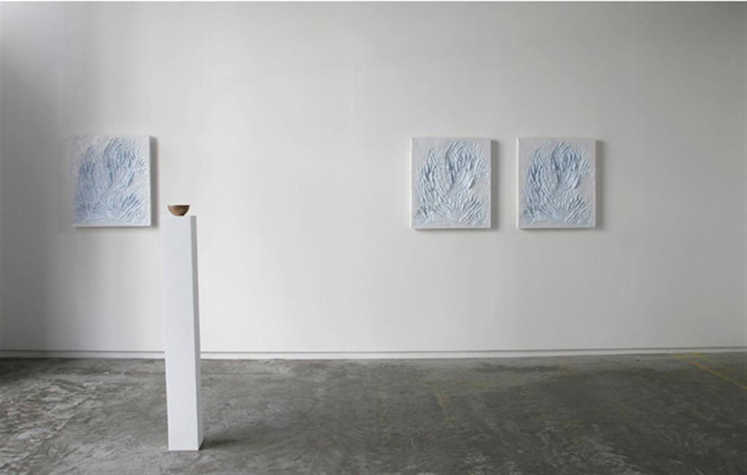 narrow white plinth with bowl atop in a white gallery space. on the walls are three white and blue textured paintings.
