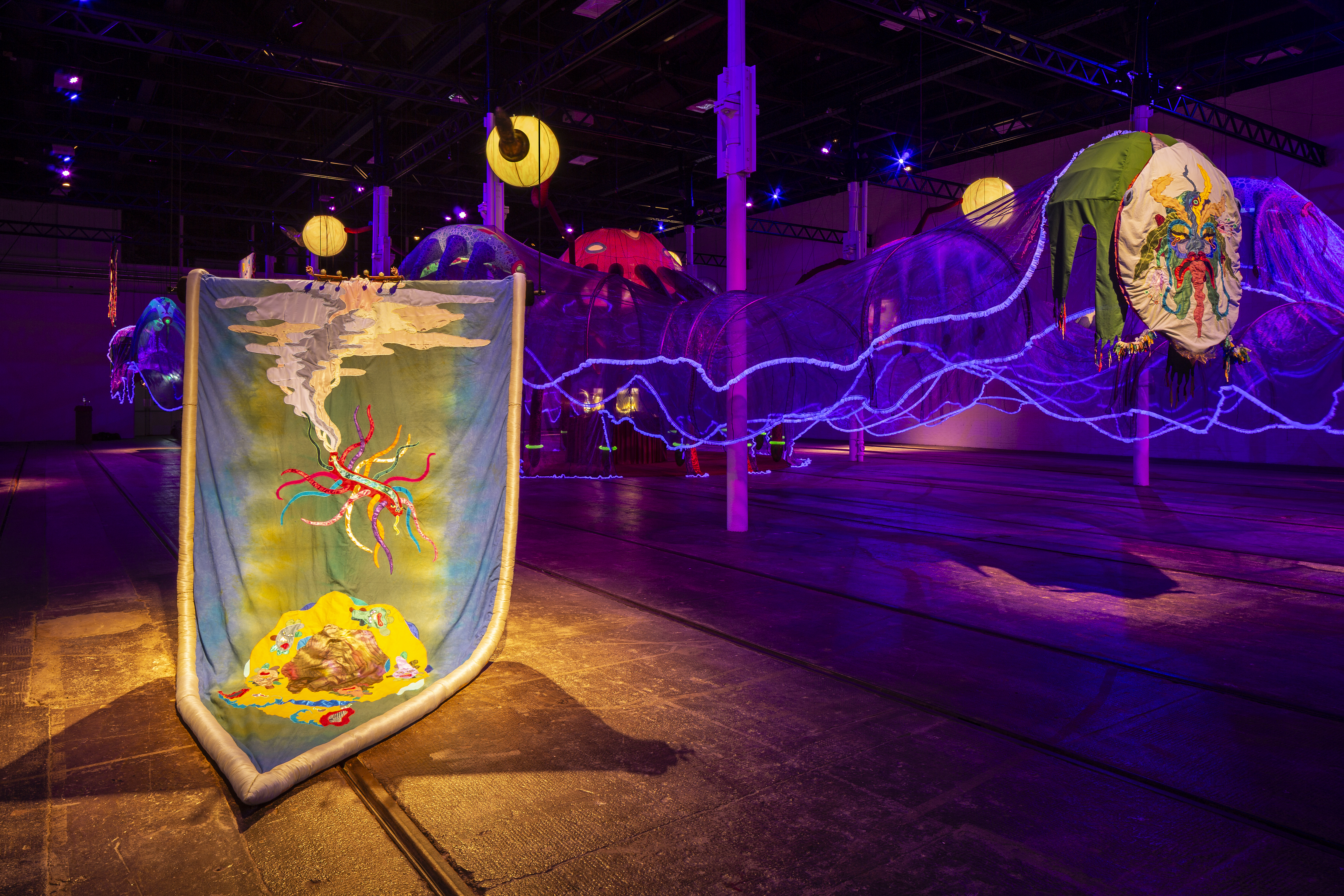 An installation shot. In the foreground is a textile work that resembles a family crest, within the crest is blue, green, and red dyed materials with embroidered flame like shapes and smoke. In the foreground of the image is the tencales of the octopuss installation. The room is dark and lit with purple and yellow hues. 