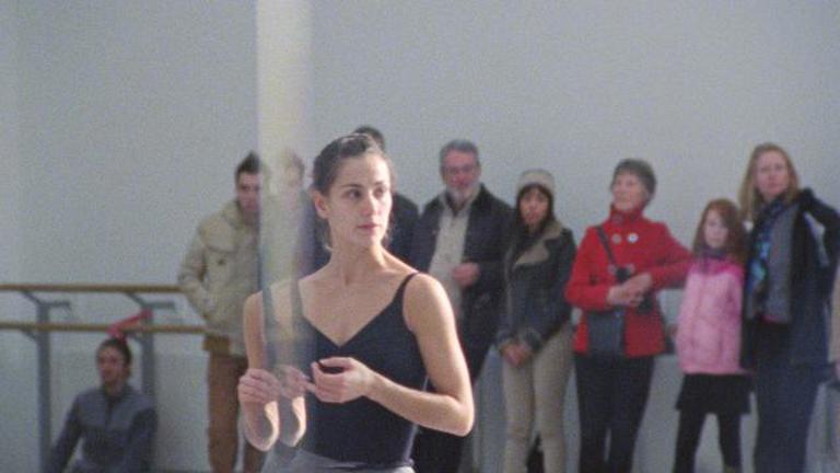 film still showing a dancer in a studio with a small crowd of spectators behind her.