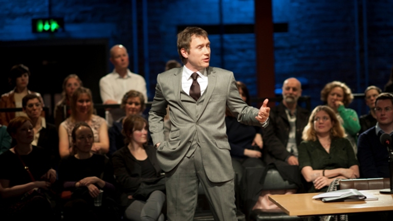 man wearing a grey three piece suit standing in front of an audience