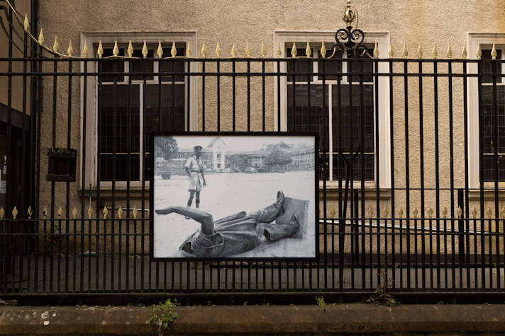 a billboard displays an intricate pencil drawing of a toppled, headless statue and a uniformed person looking over it