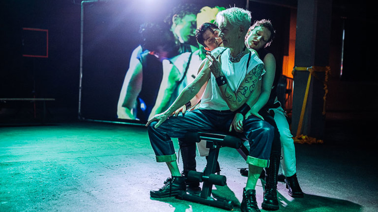 Three performers cluster together on a bench under green lighting, an older performer with grey cropped hair in the centre, with a projected image of the group behind them.