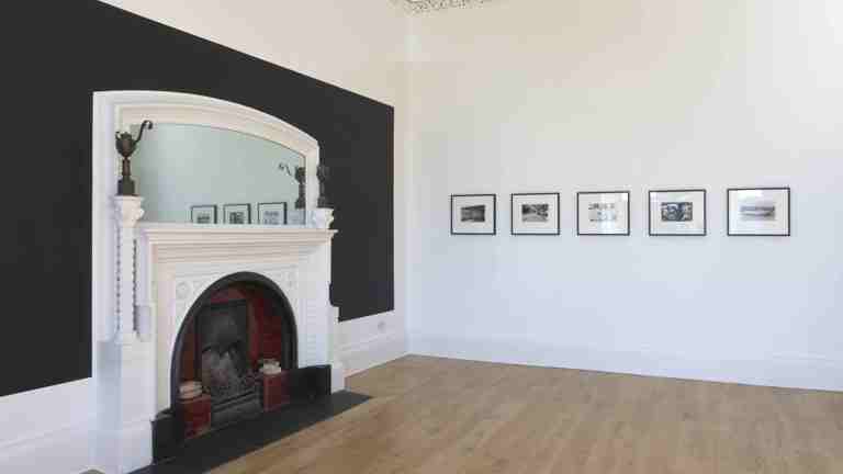 room with a white fireplace and mirror on one wall. on the other wall are five images in black frames.