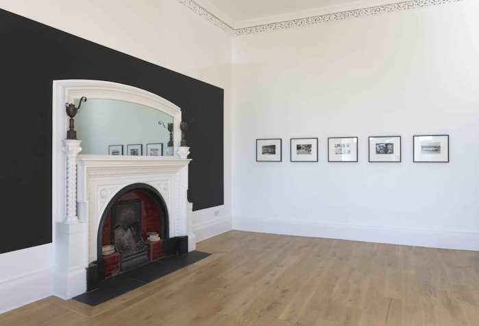 room with a white fireplace and mirror on one wall. on the other wall are five images in black frames.