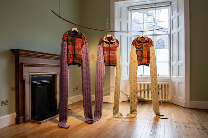 suspended on a curved wire frame hung from ceiling knitted cardigans with extra long arms, in a room with wooden flooring, fireplace, wooden sash windows and dark green walls