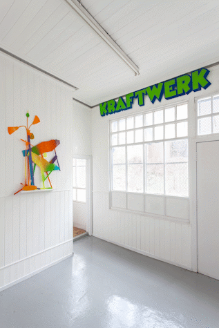 In the corner of a room, there is a sculpture hanging on a wall on the left and the word "Kraftwerk" stuck on the opposite wall, above a large window.