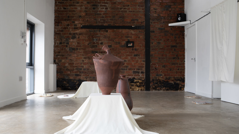 A large, urn-shaped terracotta ceramic sculpture with looping handles sits on a white draped plinth against an exposed brick wall, with smaller vessels and framed artworks arranged around the gallery space.