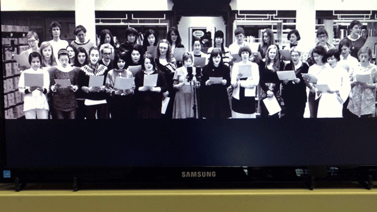 A library book shelf with a television monitor on top showing a still black and white image of a group of women in a choir , in a library