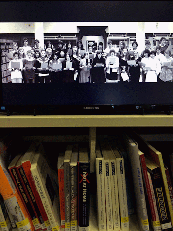 A library book shelf with a television monitor on top showing a still black and white image of a group of women in a choir , in a library