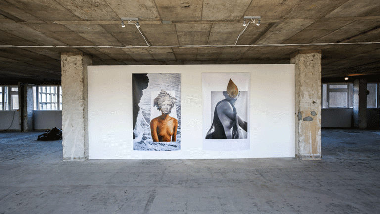 A room with concrete floors and columns has a white wall with two portraits. They mix color photographs with black and white photographs, creating a collage.