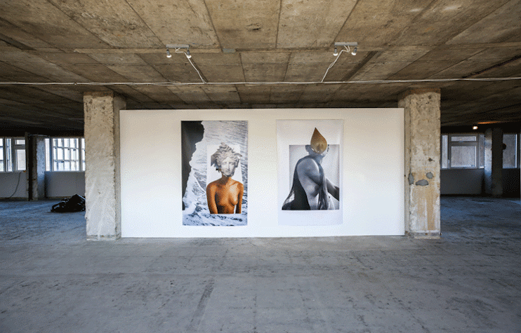 A room with concrete floors and columns has a white wall with two portraits. They mix color photographs with black and white photographs, creating a collage.