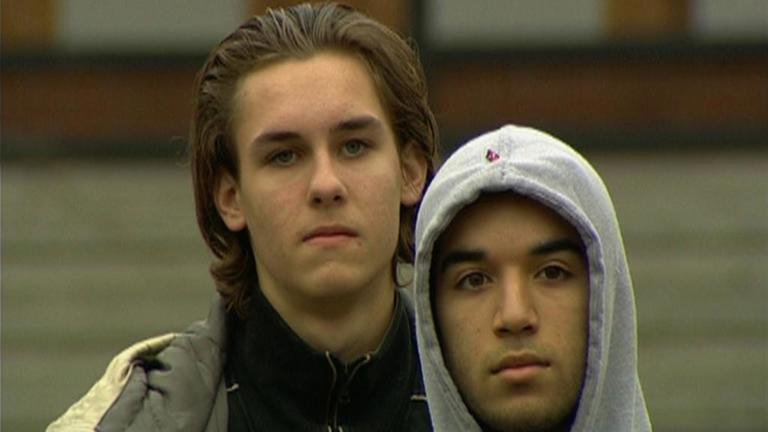 film still of two people stood looking at the camera. one wears a grey hoodie 