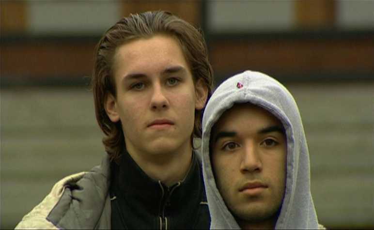 film still of two people stood looking at the camera. one wears a grey hoodie