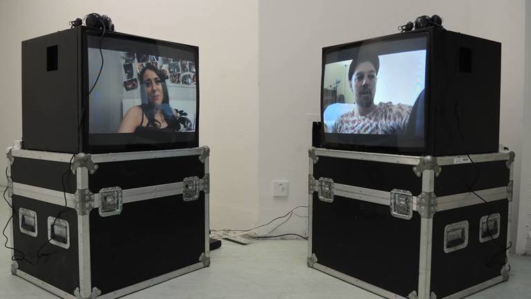 Two cube monitors on flight cases with headphones on top. On each screen the head and shoulders of a figure can be seen.  These are shown in the corner of a white room