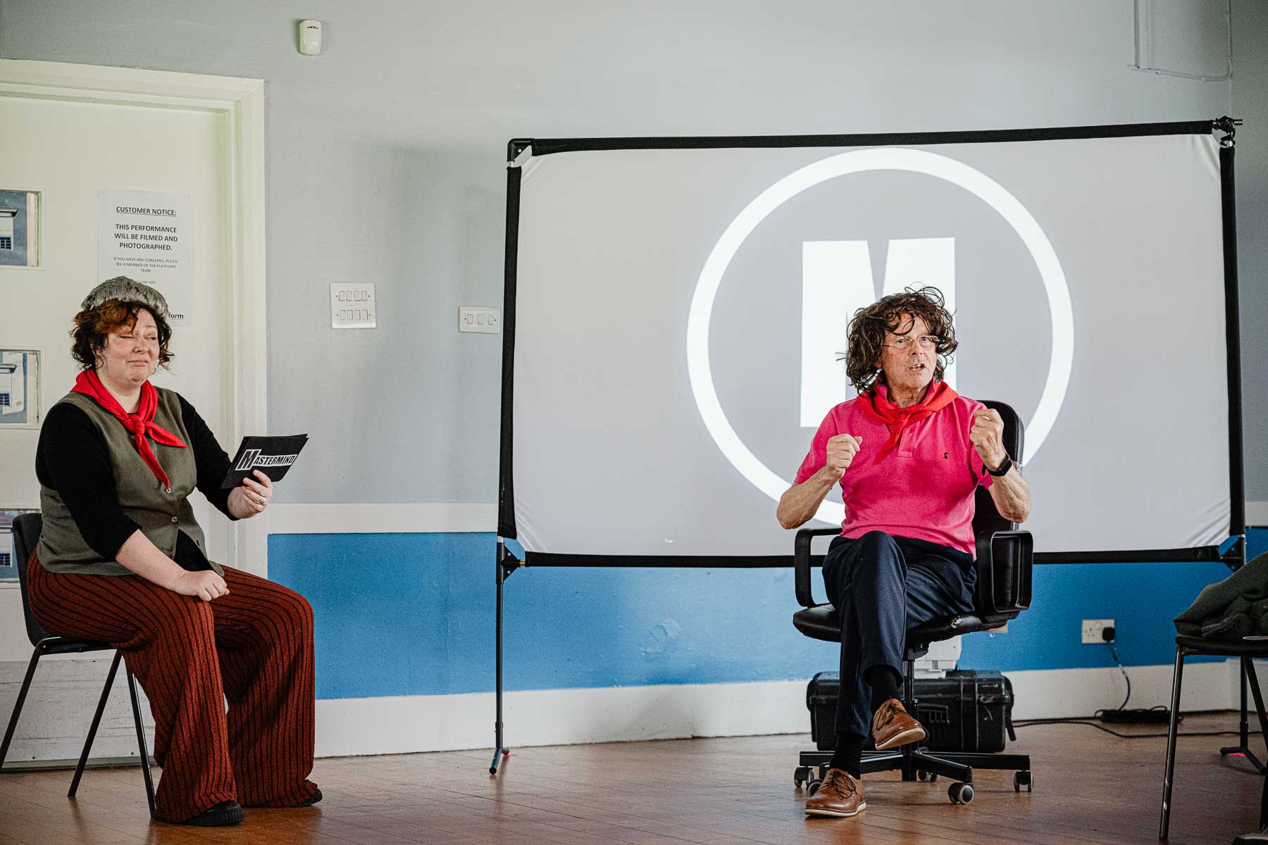 Two performers act out a mock game show scene, one holding a card reading "Mastermind," the other seated in an office chair against a projected circular logo.