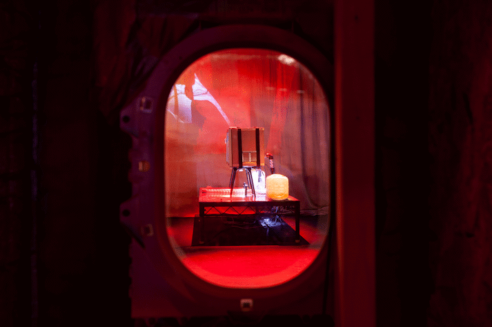 Framed by an aeroplane window, Jenkin van Zyl's installation sits in eerie red light. There is a fridge, water-tank and strange pipework.