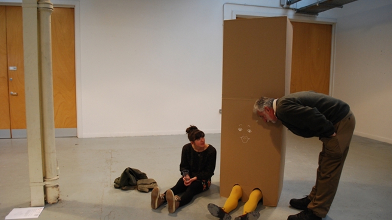 tall cardboard box with someones legs poking out the bottom. the box has a face drawn on it. next to the box is a woman sat on the floor, and on the other side of the box a man leans down from the waist as if he's talking to the box.