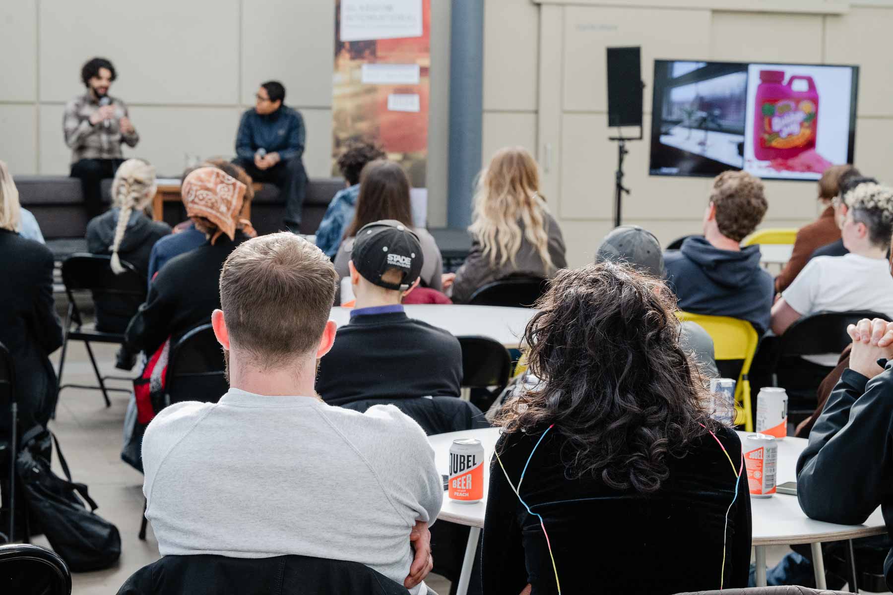 Audience members seen from behind, seated at round tables outdoors, watching two men in conversation on a stage in the background. A screen beside the stage displays an image alongside a photograph of a bottle of pink drink.