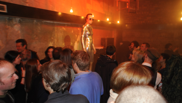 Indoor party with a crowd; person in a metallic bodysuit and sunglasses stands elevated, surrounded by dim red-orange lighting and exposed brick walls.