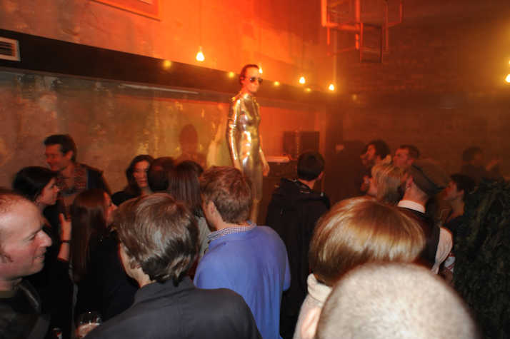 Indoor party with a crowd; person in a metallic bodysuit and sunglasses stands elevated, surrounded by dim red-orange lighting and exposed brick walls.