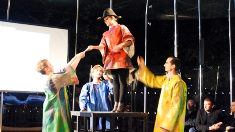 4 people in robes of green, blue, yellow, and red. the person in red stands on a crate whilst there others look up to them.