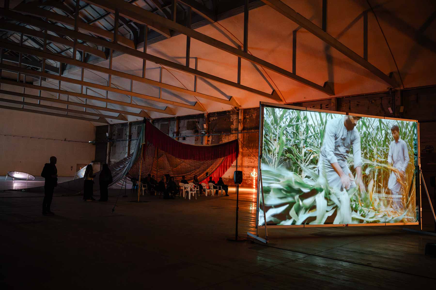 Installation view of a warehouse-style gallery with a large screen showing figures working in a maize field, red drapery and rows of chairs visible in the dimly lit space beyond.