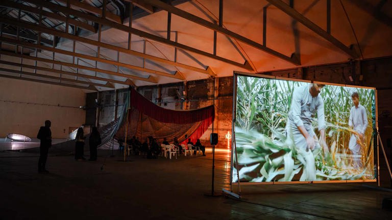 Installation view of a warehouse-style gallery with a large screen showing figures working in a maize field, red drapery and rows of chairs visible in the dimly lit space beyond.