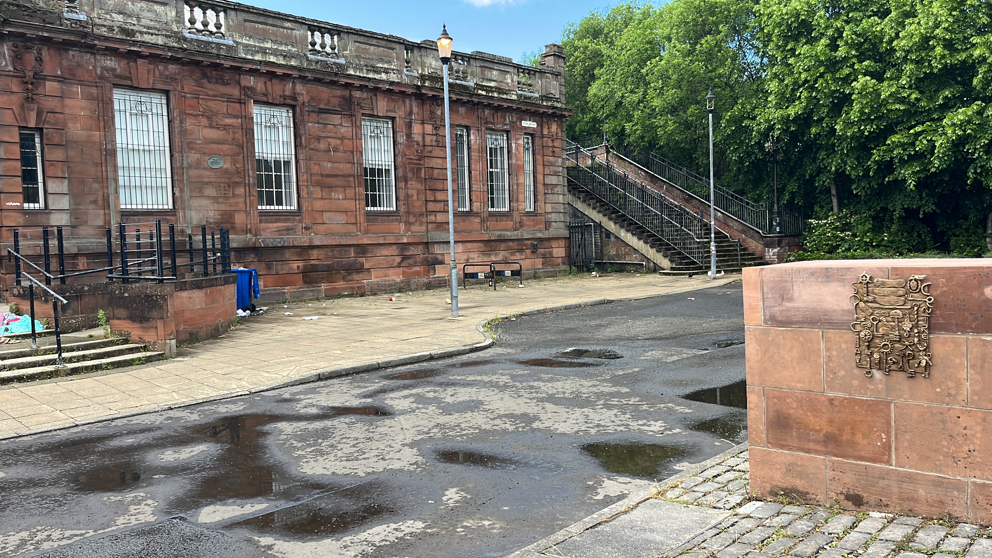 A weathered red sandstone building with barred windows and a stone staircase, beside Mandy McIntosh's stone plinth featuring a bronze relief of keys and small objects, in Springburn.