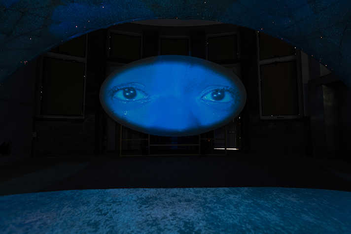 A film is projected on a dark wall. The still is a close up of a person's eyes, bathed in blue light.