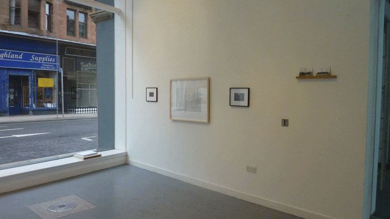 three framed works on a gallery wall and two small postcards on a wooden floating shelf. to the left is a large glass window showing the street outside.