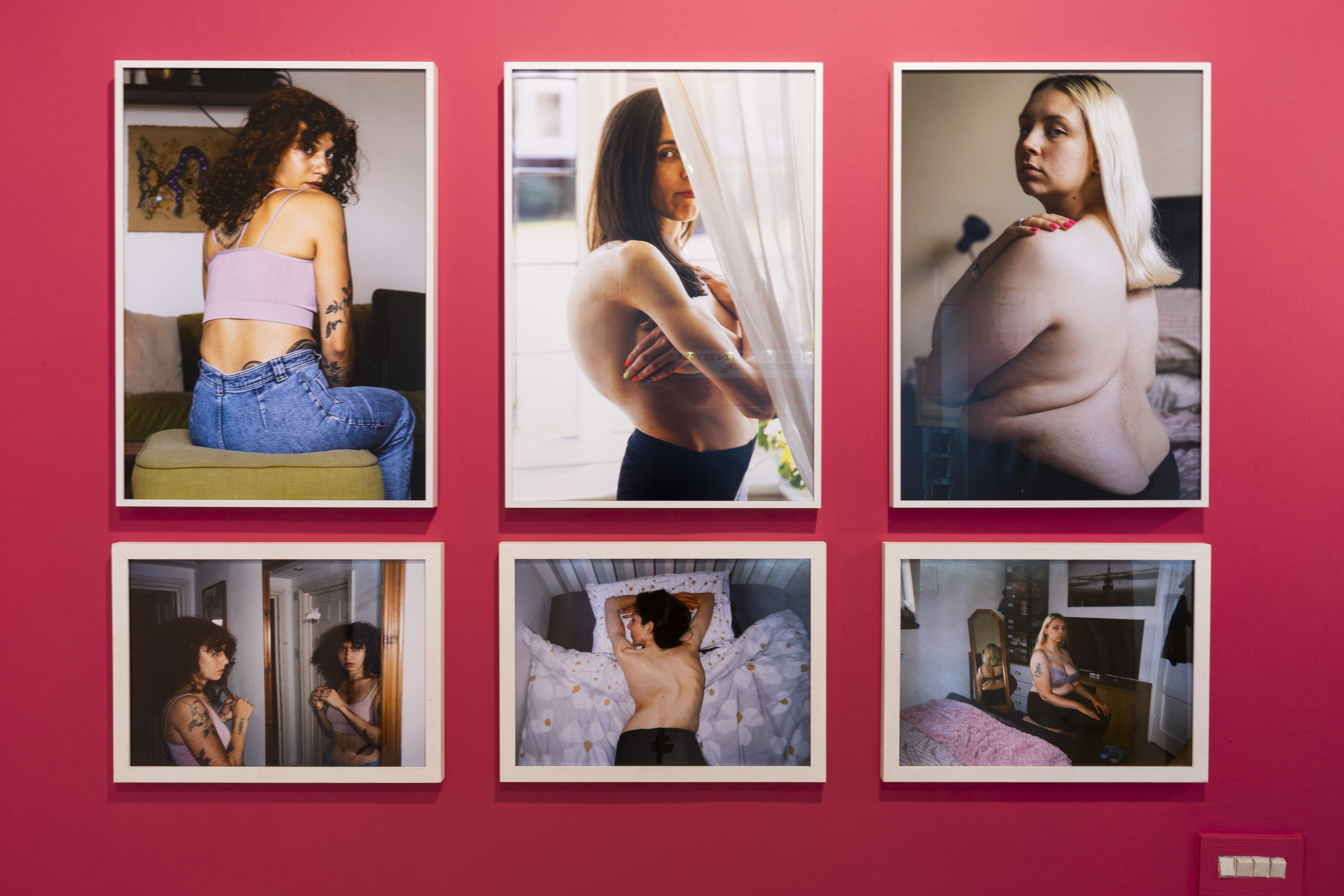 Six framed colour portraits on a pink wall showing women's bare backs and torsos in domestic settings, some with visible scars, part of Ayesha Jones' exhibition on scoliosis.