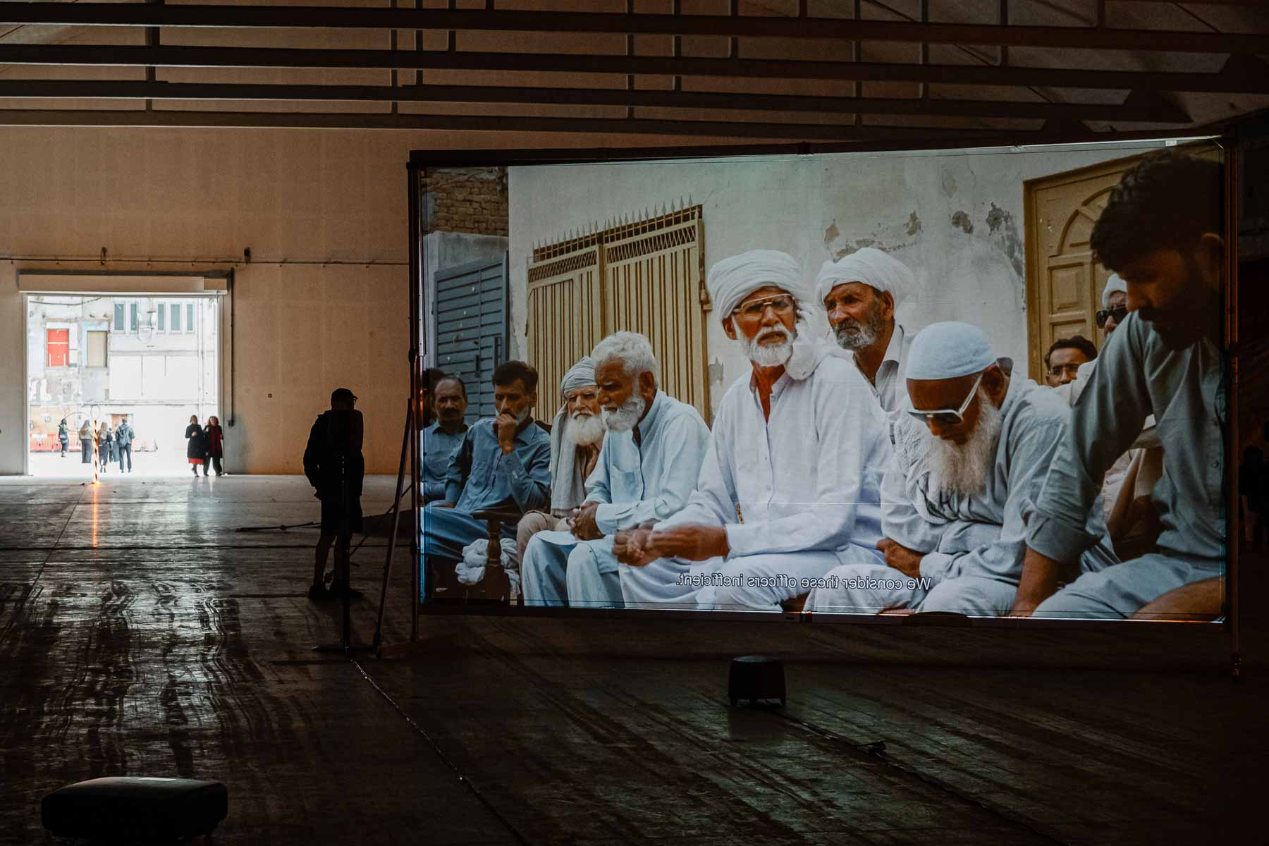 Video installation showing a group of seated people in white and grey robes with subtitled text "We consider these officers inefficient," displayed on a large screen in a warehouse-style gallery.