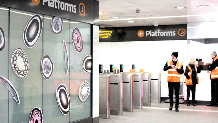 The subway entrance of Ibrox has circular drawn figures in the wall, in black, white and pink colours. Three subway station workers in their uniforms and orange vests are standing around the ticket entrance.